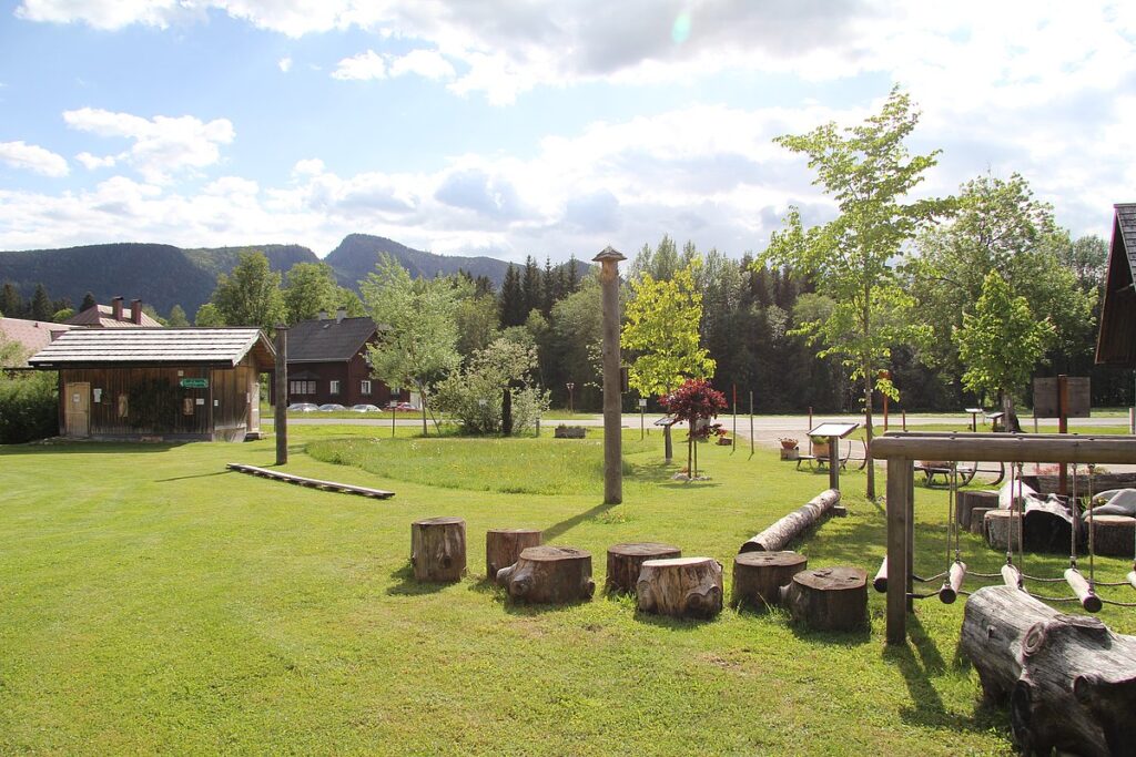 Public park scene with a grassy field, wooden play structures, and a small cabin-style building under a blue sky with clouds, mountains in the distance.