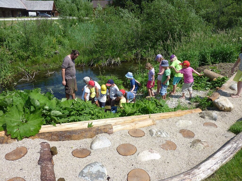 Teacher supervising a line of children by a pond in a nature garden setting.