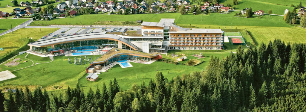 Aerial view of a modern resort with a curved glass wing, outdoor pool, and lawned grounds surrounded by a village and forest.”,